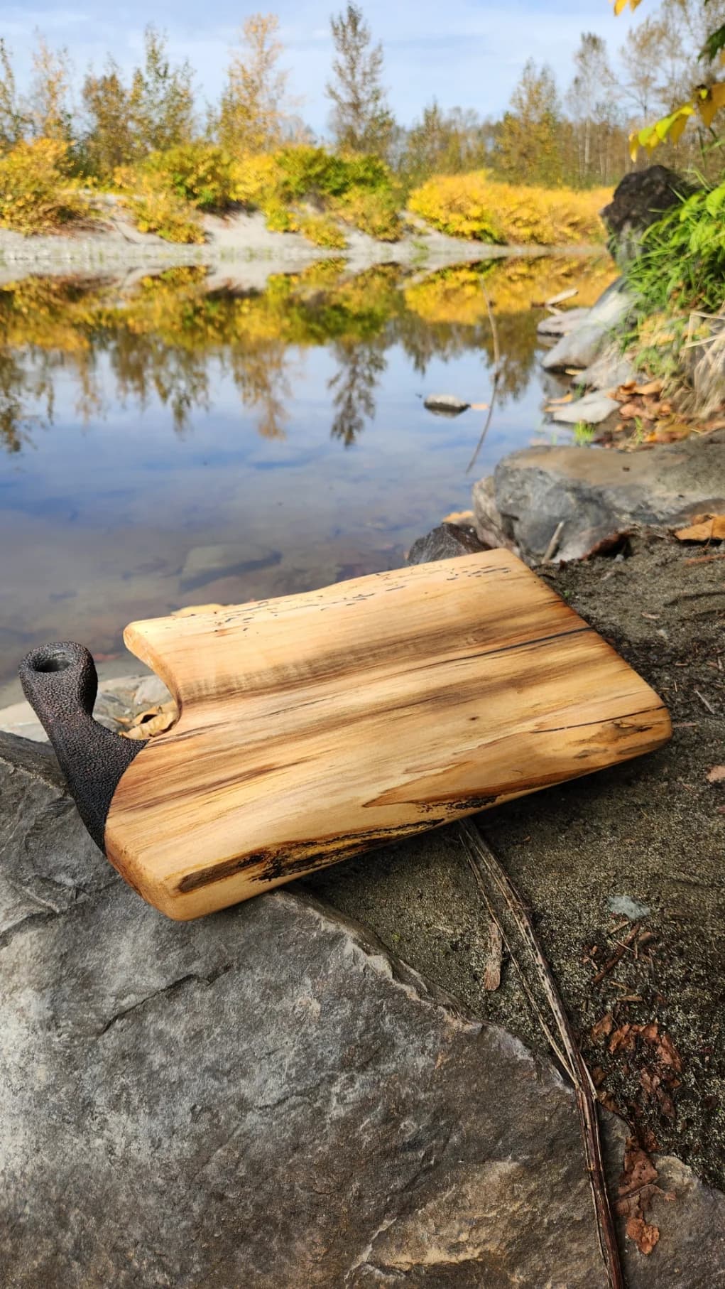 Pale live edge serving board with a dark textured handle, resting on a riverside rock in autumn